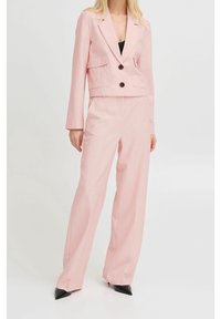 Světle růžový na míru šitý kostým s krátkým sakem se dvěma kapsami a černými knoflíky, doplněný o kalhoty s vysokým pasem a širokými nohavicemi. Černé špičaté boty.