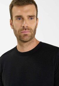 Uomo con capelli corti castani e barba, che indossa un maglione a collo tondo nero, mentre guarda leggermente di lato su uno sfondo chiaro e semplice.