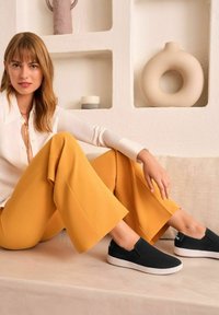 Schwarze Slipper mit strukturiertem Obermaterial, weißen Sohlen, getragen mit lockeren gelben Hosen und einer weißen Bluse, vor einem neutralen Hintergrund.