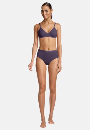 Woman standing barefoot wearing a matching dark purple wireless bra and high-waisted underwear against a plain light background.