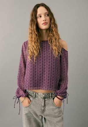 Young woman with long wavy hair wearing a purple cold-shoulder blouse and light gray high-waisted jeans, hands in pockets.