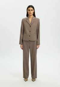 Costume marron sur mesure comprenant une veste ajustée à trois boutons, des revers en pointe et un pantalon à jambes larges. Texture lisse, motif minimal.