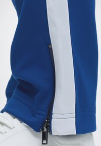 Pantalon bleu avec bande blanche sur le côté et fermeture éclair noire à la cheville, porté sur une basket blanche. Gros plan sur le bas de la jambe et la chaussure.