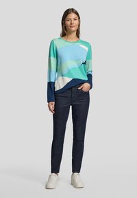 Mehrfarbiger Strickpullover mit blauen, grünen und weißen Wellenmustern. Kombiniert mit dunkler Jeans und weißen Sneakers.