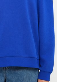 Pulover albastru regal cu manșete și tiv dublu; realizat dintr-un material moale. Purtați peste blugi din denim albastru deschis. Detaliile cusăturii sunt vizibile.