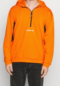 Orange hoodie med frontdragkedja, svarta sidpaneler, kenguruficka och Adidas-logga på bröstet. Mjuk tygkvalitet och avslappnad passform.