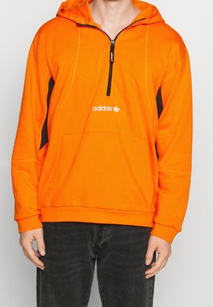 Felpa arancione con zip frontale, pannelli laterali neri, tasca a marsupio e logo Adidas sul petto. Tessuto morbido e vestibilità rilassata.