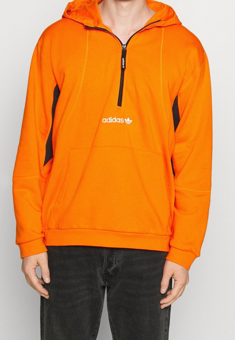 Orange hoodie med frontdragkedja, svarta sidpaneler, kenguruficka och Adidas-logga på bröstet. Mjuk tygkvalitet och avslappnad passform.