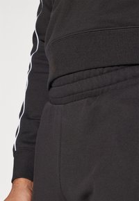 Czarny sweatshirt i dresy z białymi bocznymi paskami. Miękki, fakturowany materiał z prążkowanymi mankietami i detalami w pasie. Ujęcie z bliska.