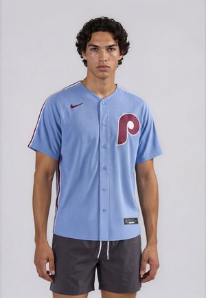 Joven con pelo rizado que lleva una camiseta de béisbol azul con un logo "P" color burdeos y pantalones cortos oscuros sobre un fondo liso.