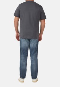 Man die met zijn rug naar de camera staat, gekleed in een donkergrijze t-shirt, blauwe versleten jeans en witte sneakers tegen een effen witte achtergrond.