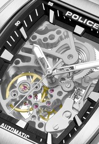 Srebrny zegarek mechaniczny z przezroczystą tarczą. Posiada skomplikowane przekładnie, złote akcenty, purpurowe klejnoty oraz czarną obręcz z białymi oznaczeniami.