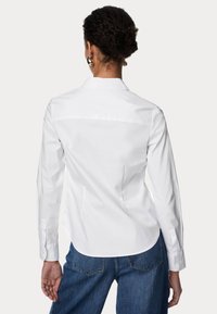 Hvid skjorte med knapper og klassisk krave, lange ærmer og rygdetalje, parret med blå jeans. Glat bomuldsstof, uden mønstre.