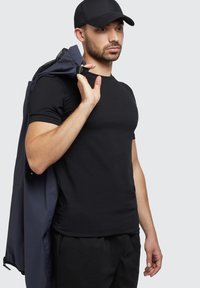 Fekete, kerek nyakú póló, testhezálló fazonban, egy sötétkék dzsekit viselve a vállán. A modellen sötét haj található és fekete sapkát visel.