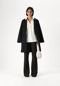 Black oversized coat over a white button-up shirt, paired with dark green textured trousers. Holding a light beige bag with a logo.