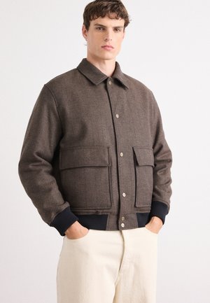 Brown herringbone textured jacket with a collared neck, two front patch pockets, snap buttons, and navy ribbed cuffs and hem.