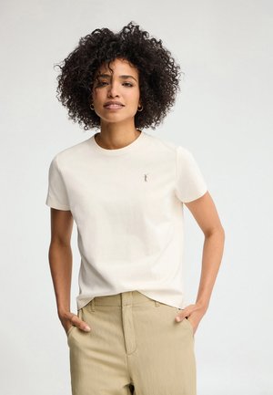 Kobieta o kręconych włosach stoi uśmiechnięta, ubrana w prostą białą koszulkę z krótkim rękawem i jasnobeżowe spodnie, dłonie w kieszeniach, neutralne tło.