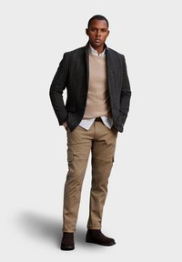 Completo da uomo con un blazer a righe verticali scure, maglione beige, camicia bianca e pantaloni cargo color khaki, abbinato a stivali alla caviglia marroni.