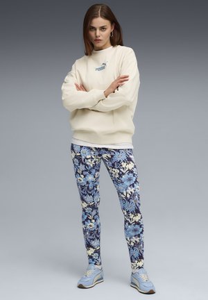 Felpa crema con logo discreto, abbinata a leggings blu con motivo floreale e dettagli bianchi. Sneakers azzurre completano il look.