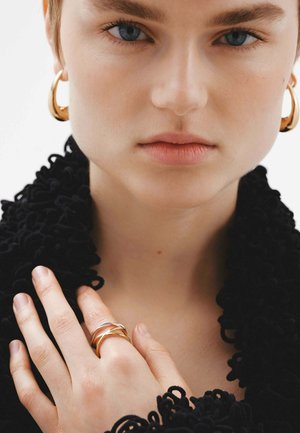 Pendientes de aro dorados y anillos entrelazados en los dedos, con una prenda negra texturizada que cae sobre los hombros. Tonos de piel suaves y naturales.