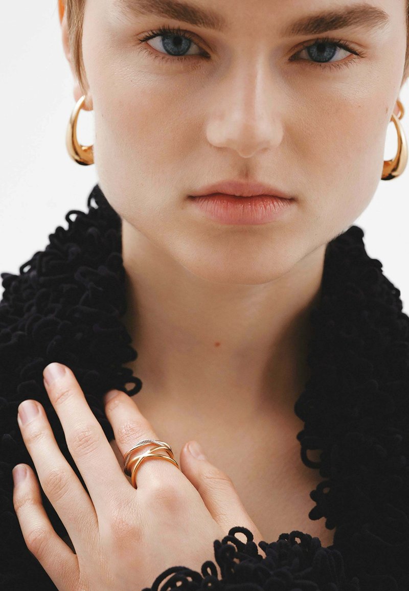 Pendientes de aro dorados y anillos entrelazados en los dedos, con una prenda negra texturizada que cae sobre los hombros. Tonos de piel suaves y naturales.