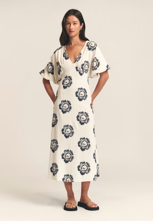 Robe blanche avec un motif floral bleu marine et rouille, à manches larges et courtes, décolleté en V profond et coupe décontractée, associée à des sandales plates noires.