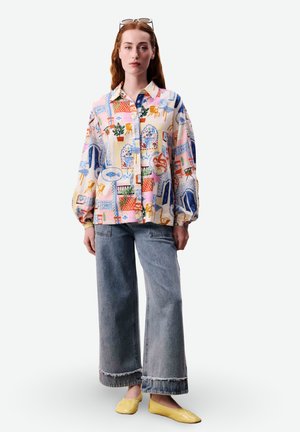 Femme portant une chemise boutonnée oversize colorée à motifs, un jean large délavé avec des ourlets bruts, des ballerines jaunes et des lunettes de soleil sur la tête.