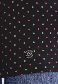 Czarny materiał w różowe i szare kropki, mała okrągła czarna metka z wzorem serca, a poniżej niebieski denim.