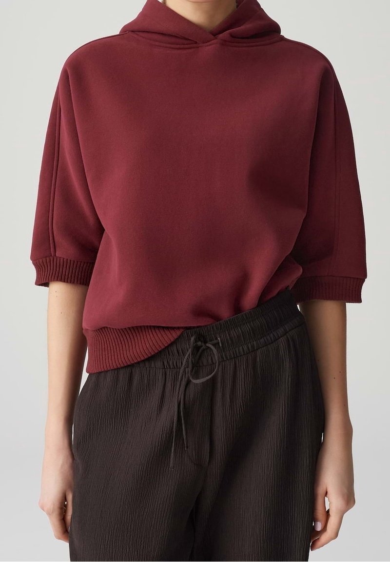 Personne portant un sweat à capuche bordeaux à manches courtes et un pantalon noir texturé avec cordon, debout devant un fond clair uni.