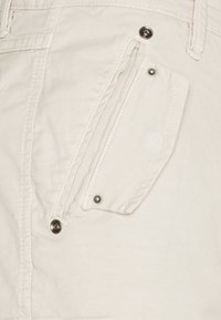 Bílé bavlněné kalhoty s boční kapsou s klopou, uzavíratelnou stříbrnými patentkami, a texturovanou tkaninou s jemnými vertikálními pruhy.