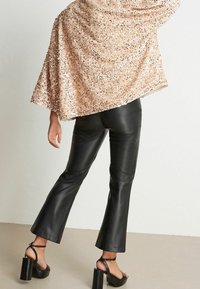 Cape en or rose pailleté, portée sur un pantalon évasé en similicuir noir, associée à des sandales noires à talons blocs. Textures lisses et détails brillants.