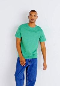Groene katoenen t-shirt met ronde hals en korte mouwen, met een subtiel logo op de borst, gecombineerd met blauwe denim jeans. Effen achtergrond.