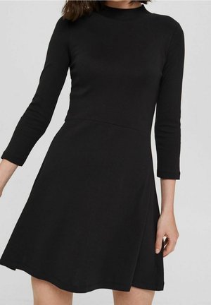 Femme portant une robe noire à manches longues, longue jusqu'aux genoux, avec une taille cintrée et un col rond, debout sur un fond clair.
