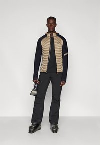 Beige gewatteerd jack met marineblauwe mouwen, zwarte broek en skischoenen. Houdt een skipas vast. Heeft een ritssluiting aan de voorkant en oranje accenten.