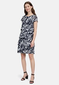 Granatowa sukienka z białymi wzorami liści, krótkimi rękawami, falbankowym wzorem w warstwy i okrągłym dekoltem, zestawiona z czarnymi sandałami na obcasie.
