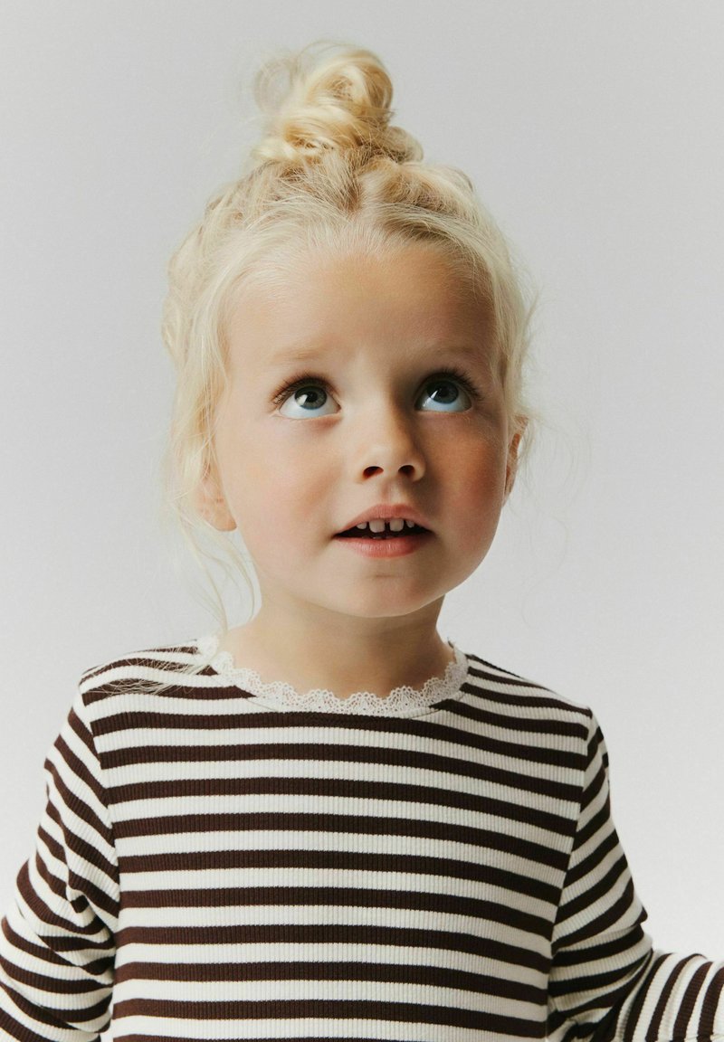 Jeune fille blonde avec un chignon en haut de la tête, portant un t-shirt rayé noir et blanc, regardant vers le haut avec une expression curieuse sur un fond uni.