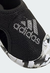 Scarpa da ginnastica nera in mesh con logo bianco, dotata di una suola in camouflage nera e grigia con texture. Progettata per comfort e flessibilità.