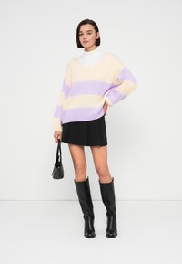 Pull à col en V, oversized, crème avec des rayures lavande et violet clair. Associé à une jupe plissée noire et des bottes noires mi-mollet.