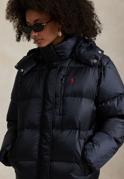 Vrouw met krullend haar die zwarte zonnebril draagt en een marineblauwe pufferjas met een rood polo-logo en schelpvormige oorbellen