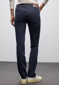 Persona con jeans de un azul oscuro y zapatillas blancas de pie sobre un suelo gris, contra una pared gris clara.