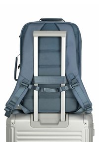 Mochila con correas acolchadas ajustables, fabricada en material texturizado azul oscuro. Cuenta con un asa de aluminio gris situada sobre una maleta rodante plateada.