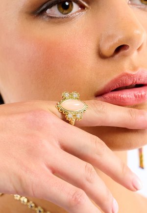 Gold ring featuring an oval pale stone, surrounded by clear gemstones, set on a textured band, showcased on a hand near a face.