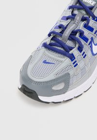 Nike tenisice u sivoj i plavoj boji, s mrežastim gornjim dijelom, podstavljenim jezikom i gumenim potplatom s istaknutim logom i vezicama.
