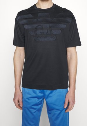 Mand iført en sort T-shirt med korte ærmer med præget GA-logo og blå bukser, stående mod en ensfarvet hvid baggrund.