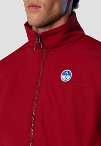 Giacca rossa con colletto alto, zip frontale e un logo circolare blu e bianco sul petto. Tessuto liscio e design minimalista.