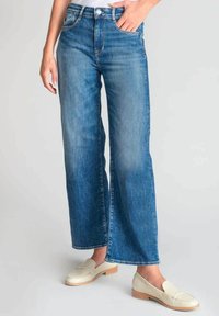 Jean bleu à jambes larges avec un délavage, taille haute et un design classique à cinq poches, associé à des mocassins crème.