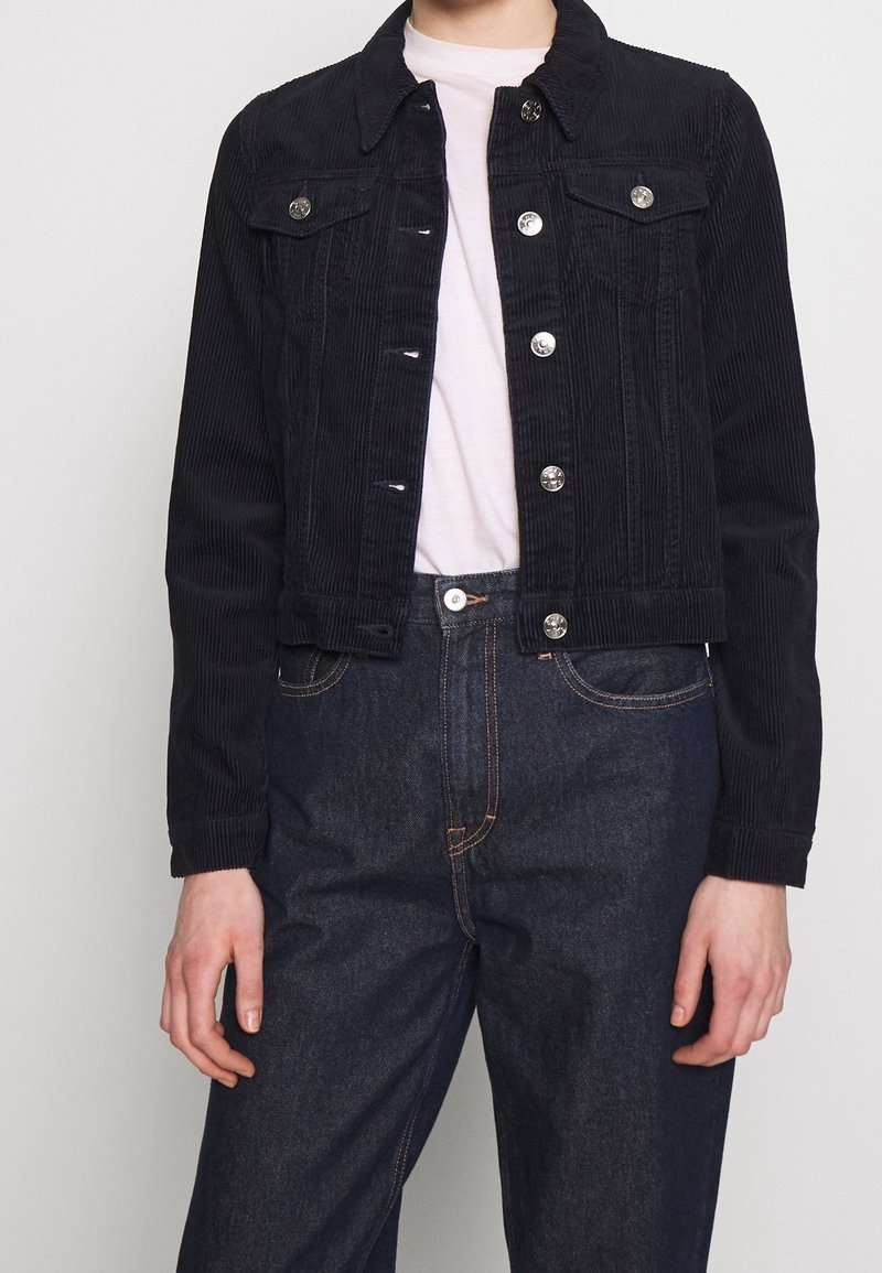 Personne portant une veste en velours côtelé noire avec des boutons métalliques sur une chemise blanche et un jean denim bleu foncé sur un fond uni.