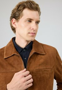 Bruine suede jack met een ritssluiting aan de voorkant, scherpe kraag en een borstzak. Gedragen over een marineblauw overhemd met knopen, met een gladde textuur.