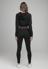 Hoodie noir avec poche kangourou, associé à un jogging noir ajusté. Présente une ceinture de marque SIKSIL avec des accents roses. Baskets blanches.