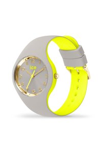 Orologio in silicone grigio con cinturino giallo; il quadrante rotondo presenta dettagli dorati, un quadrante grigio e una manina gialla evidente; design minimalista.
